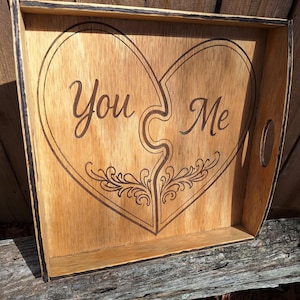 May include: Wooden serving tray with a heart design. The tray is made of light-colored wood and features the words "You" and "Me" within a heart-shaped design. The tray has handles and a decorative floral element.