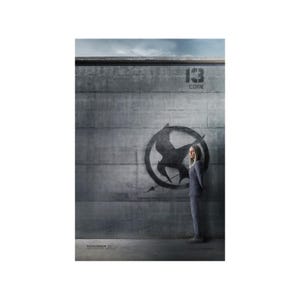 May include: A woman in a gray suit stands in front of a concrete wall with a large gray and black mockingjay symbol painted on it. The text "13 COIN" is above the symbol. The text "NOVEMBER 21" is at the bottom of the image.