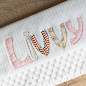May include: White baby blanket with the name "Livvy" embroidered in pink, brown, and beige fabric letters.