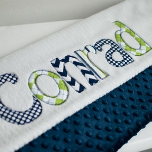 May include: A white and blue baby blanket with the name "Conrad" embroidered on it in green, white, and blue fabric. The blanket has a blue minky backing.