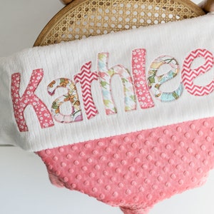 May include: A white and pink baby blanket with the name "Kathleen" embroidered on it in colorful fabric letters.