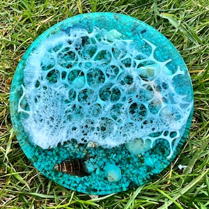 Puede incluir: Un posavasos redondo de resina azul turquesa con remolinos de resina blanca que se asemejan a olas. El posavasos está lleno de pequeñas piedras y conchas azules turquesas y blancas.