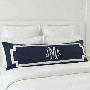 May include: A long, rectangular navy blue lumbar pillow featuring a white border frame and the white embroidered monogram letters JMK centered on the front. The pillow is positioned horizontally across a bed with white bedding and pillows.