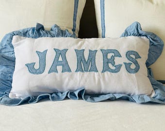 Op de afbeelding: Een wit kussen met blauwe denimbies en ruches. Het kussen heeft de naam "JAMES" geborduurd in blauwe draad.