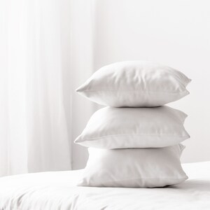 May include: Three white pillows stacked on top of each other on a white bed.