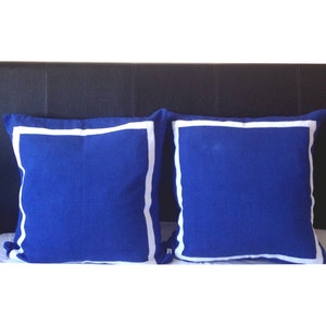 May include: Two blue throw pillows with white piping on a black leather headboard. The pillows are square and have a soft, plush texture.