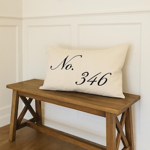 May include: A wooden bench with an off-white pillow that has the text "No. 346" in black script. Above the bench, mounted on the white wall, is a black metal coat rack with four hooks. The bench is made of wood with an X-shaped design on the sides.