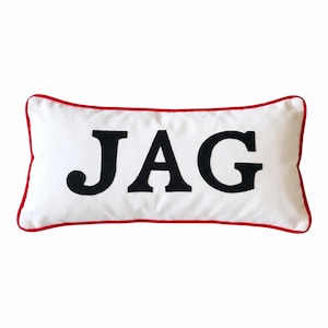 May include: A rectangular white throw pillow featuring the black capital letters JAG centered on the front. The pillow is finished with a thin red piped trim along all four edges, creating a distinct border against the white fabric background.
