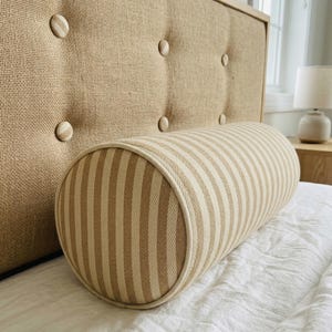 Op de afbeelding: Een beige gestoffeerd hoofdbord met knopen en een gestreept bolsterkussen. Het bolsterkussen heeft verticale beige en witte strepen en een witte bies. Het hoofdbord is gemaakt van een geweven stof.