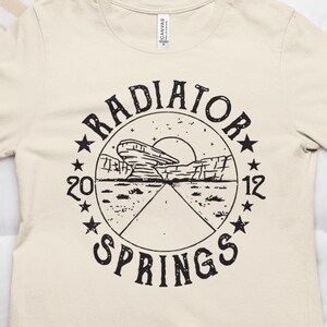May include: A beige t-shirt with a black graphic print of a circle with the text "RADIATOR SPRINGS" and a stylized image of a car driving through a desert landscape.