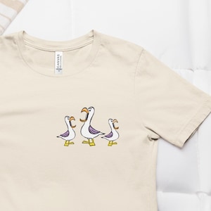 May include: A cream-colored t-shirt featuring a cartoon design of three seagulls with open beaks. The seagulls have white bodies, purple wings, and yellow feet. The shirt is made of a soft material and has a classic crew neck.