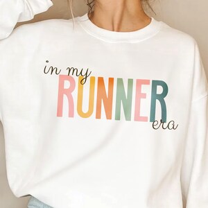 May include: White sweatshirt with the text "in my runner era" in rainbow colors.