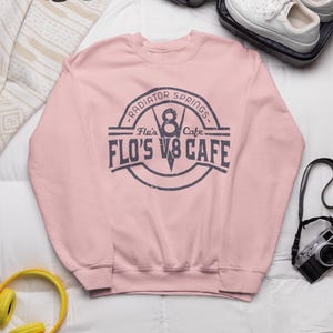 May include: Pink sweatshirt with a vintage-style logo that says "Flo's V8 Cafe - Radiator Springs - Flo's Cafe".