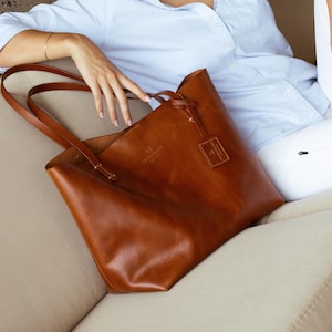 May include: A brown leather tote bag with a long strap and a small leather tag that says "The  Ancher." The bag is sitting on a light brown couch.