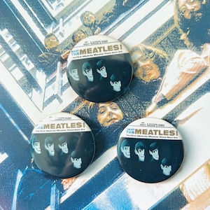 Puede incluir: Tres botones redondos en blanco y negro con el texto "THE MEATLES!" y la frase "The First Album by Whoever These Guys Are." Cada botón presenta una imagen estilizada de cuatro hombres.