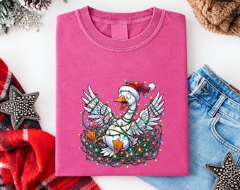 May include: A pink t-shirt featuring a graphic of a white goose wearing a red Santa hat and tangled in a string of colorful Christmas lights. The goose has an open beak and wide eyes, with its wings spread out amidst the glowing light bulbs.