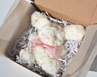 Op de afbeelding: Een witte pluche konijn met roze oren en een roze strik in een bruine kartonnen doos met wit tissuepapier.
