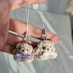 Könnte beinhalten: Zwei Plüsch-Teddybär-Schlüsselanhänger mit weißem Fell und gestickten Gesichtern. Ein Bär hat eine lila Schleife und der andere eine weiße Schleife.