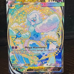 Può includere: Una carta Pokémon Mega Altaria EX con un design colorato. La carta presenta una creatura simile a una nuvola nei toni del blu e del bianco, con dettagli dorati e il testo "Mega Harmony". La carta è esposta su un supporto trasparente.