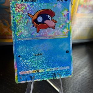 Op de afbeelding: Een holografische Pokémon-verzamelkaart met het personage Shellder. De kaart heeft een blauwe achtergrond met het personage in het midden, een rode en oranje schelp met een donkerblauw lichaam en een roze tong. De kaart wordt weergegeven op een heldere acrylstandaard.
