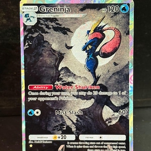 May include: A holographic Greninja Pokémon card. The card features a blue and red Greninja character with the text "Greninja" and "Water Shuriken". The card has a HP of 120 and the text "Mist Slash".