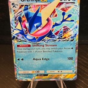 May include: A Greninja ex Pokémon trading card, featuring a blue and pink character with a yellow and white accent. The card displays the text "Greninja ex", "HP 170", and "Aqua Edge 100". The card is displayed on a clear plastic stand.