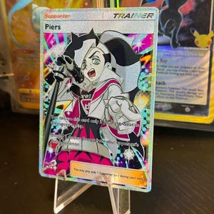 May include: A holographic "Piers" Trainer card, featuring a black and white illustrated character with a microphone. The card has text that reads "Supporter" and "Trainer". Other cards are visible in the background.