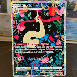 May include: A Pokémon card featuring Milotic with a HP of 120. The card has a black background with floral designs and the text "Healing Ripples" and "Aqua Edge". The card is displayed on a clear stand.