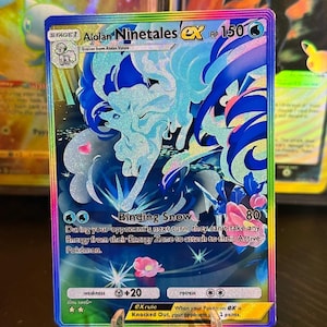Könnte beinhalten: Holografische Alolan Ninetales EX Pokémon-Sammelkarte. Die Karte zeigt eine blau-weiße Illustration des Pokémon mit dem Text "Binding Snow". Die Karte wird auf einem transparenten Ständer präsentiert.