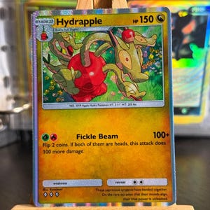 May include: A holographic Hydrapple trading card featuring a green, multi-headed creature emerging from a red apple. The card displays 150 HP, the Fickle Beam attack, and text describing the creature. The card is set on a small wooden easel.