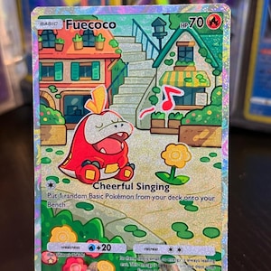 Puede incluir: Una carta coleccionable Pokémon holográfica con Fuecoco, una criatura roja y amarilla con forma de cocodrilo. La carta tiene un fondo colorido con edificios y una flor. El texto incluye "Fuecoco", "Cheerful Singing" y otros detalles del juego.