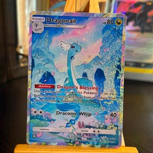 May include: A holographic Dragonair trading card featuring a blue serpentine creature with small wings in a vibrant, colourful landscape. The card displays the text Dragonair, HP 80, Dragon's Blessing ability, and Draconic Whip attack with 40 damage.