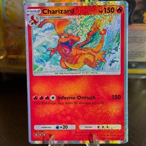 May include: A holographic Charizard Pokémon card, Stage 2, with a red and orange design. The card features the text "Charizard" and "Inferno Onrush". The card is displayed on a clear stand.