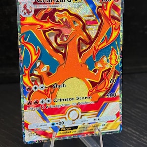 May include: A Charizard EX Pokémon trading card. The card features an orange Charizard with red flames against a blue and gold background. Text includes "Charizard EX", "Slash", and "Crimson Storm". The card is displayed on a clear plastic stand.
