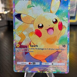 Peut inclure: Une carte à collectionner Pokémon holographique Pikachu. La carte présente un Pikachu souriant avec des joues rouges et un corps jaune. La carte porte les mentions "Pikachu", "Spark" et "HP 60". La carte est présentée sur un présentoir en plastique transparent.