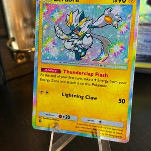 Può includere: Una carta collezionabile Pokémon olografica di Zeraora. La carta presenta uno sfondo giallo con il personaggio Zeraora, una creatura bipede bianca, blu e gialla. La carta ha il testo "HP 90" e "Lightning Claw 50".