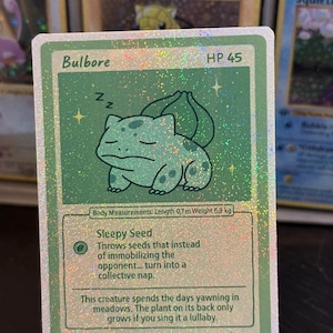 Könnte beinhalten: Eine holografische Bulbore Pokémon-Sammelkarte. Die Karte zeigt eine schlafende Bulbore-Illustration mit dem Text "Bulbore" und "HP 45". Die Karte enthält auch den Text "Sleepy Seed" und "12/50". Die Karte ist 70 cm lang und wiegt 6,9 kg.
