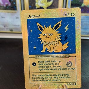 Op de afbeelding: Goudkleurige ruilkaart met het personage "Joltired" op een blauwe achtergrond met bliksem. De kaart heeft "HP 90" en beschrijft vaardigheden en afmetingen: 0,8 m lang, 24 kg.