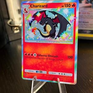 Op de afbeelding: Een holografische Charizard Pokémon-kaart, Fase 2, met een rode en oranje achtergrond. De kaart toont een illustratie van een zwarte en oranje draak met de naam "Charizard" bovenaan. De kaart wordt gepresenteerd op een transparante plastic standaard.