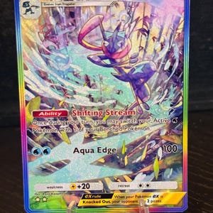 May include: A holographic Greninja EX Pokémon card with a rainbow border. The card features a blue and purple Greninja character with the text "Greninja EX", "HP 170", and "Aqua Edge 100". The card is displayed on a clear stand.