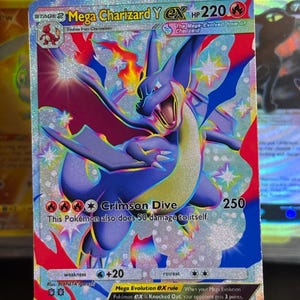 May include: A holographic Mega Charizard Y EX trading card featuring a blue dragon-like creature with red wings, 220 HP, and the Crimson Dive attack. The card displays text including Mega Charizard Y, 250 damage, and the Mega Evolution EX rule.