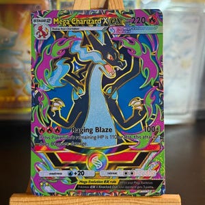 Może przedstawiać: Karta kolekcjonerska Mega Charizard X EX przedstawiająca czarnego smoka z niebieskimi płomieniami na tle żywych fioletowych i zielonych kolorów. Karta zawiera 220 HP, atak Raging Blaze oraz tekst opisujący zasadę Mega Evolution EX.
