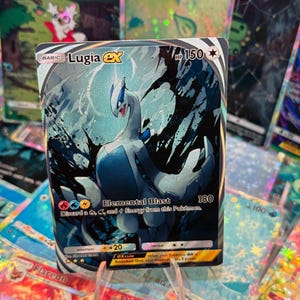 Puede incluir: Una carta coleccionable Pokémon holográfica de Lugia ex. La carta presenta una ilustración azul y blanca de Lugia con el texto "Elemental Blast". Otras cartas Pokémon son visibles en el fondo.