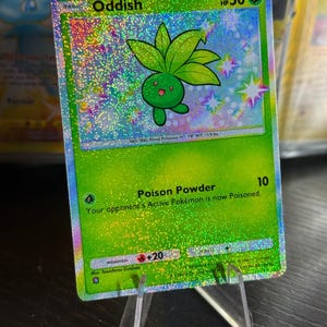 Peut inclure: Une carte à collectionner Pokémon holographique représentant Oddish, une créature verte en forme de plante. La carte a un fond vert avec le texte "Oddish" et "HP 50". La carte est présentée sur un présentoir en plastique transparent.