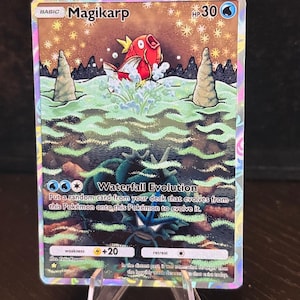 Op de afbeelding: Een holografische Magikarp Pokémon-kaart. De kaart toont een rode Magikarp die uit het water springt, met een waterval en een rotsachtig landschap op de achtergrond. Tekst bevat "Magikarp", "Waterfall Evolution" en andere speld details.
