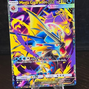 Puede incluir: Una carta coleccionable Pokémon Mega Gyarados EX. La carta presenta una vibrante ilustración de un Gyarados azul y amarillo con acentos morados. La carta muestra el texto "Mega Blaster" y "HP 210". La carta se muestra en un soporte de plástico transparente.