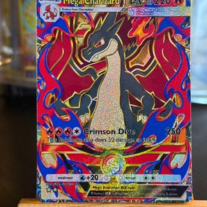 Könnte beinhalten: Eine Mega Charizard Y EX Sammelkarte mit einem schwarzen und cremefarbenen drachenartigen Wesen, umgeben von leuchtend roten und blauen Flammen. Die Karte zeigt den Text Mega Charizard Y, 220 HP, Crimson Dive und verschiedene Spielstatistiken.