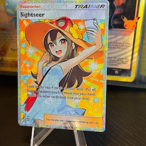 Può includere: Carta Allenatore "Sightseer" olografica del GCC Pokémon. La carta presenta un personaggio sorridente con cappello e occhiali da sole arancioni, che tiene in mano un telefono. Il testo descrive la funzione della carta.
