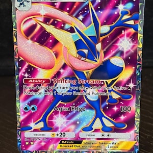 May include: A holographic Pokémon trading card featuring Greninja EX. The card displays a blue and pink ninja-like creature with the text "Greninja EX" and "HP 170". The card also includes the text "Shifting Stream" and "Aqua Edge".