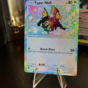 Może przedstawiać: Holograficzna karta kolekcjonerska Pokémon Type: Null. Karta zawiera kolorową ilustrację stwora, z tekstem zawierającym "Type: Null", "HP 80" i szczegóły ataku. Karta jest prezentowana na przezroczystym plastikowym stojaku.